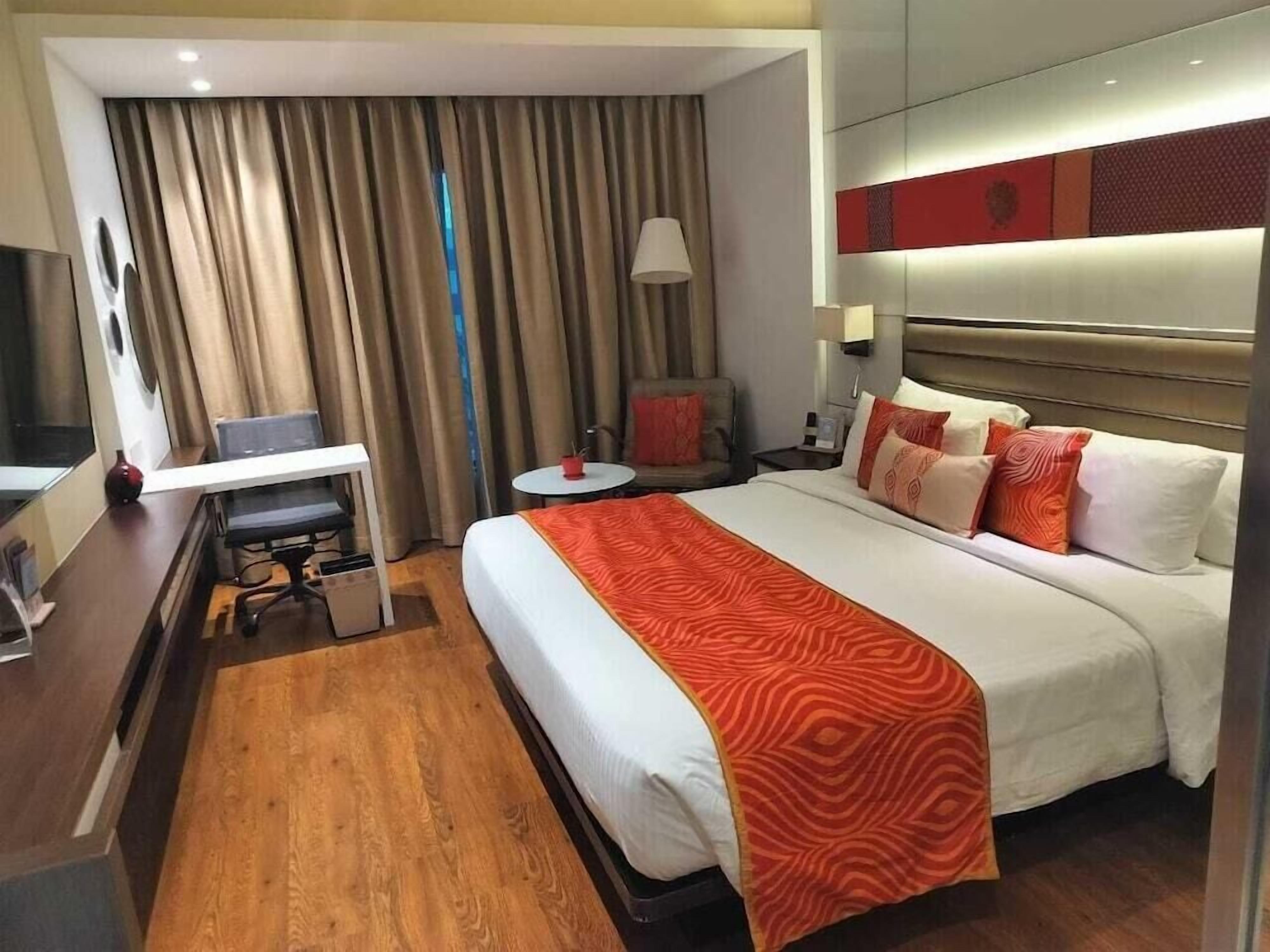 A hotel room with a bed, a desk, a chair, a small table, and a lamp.