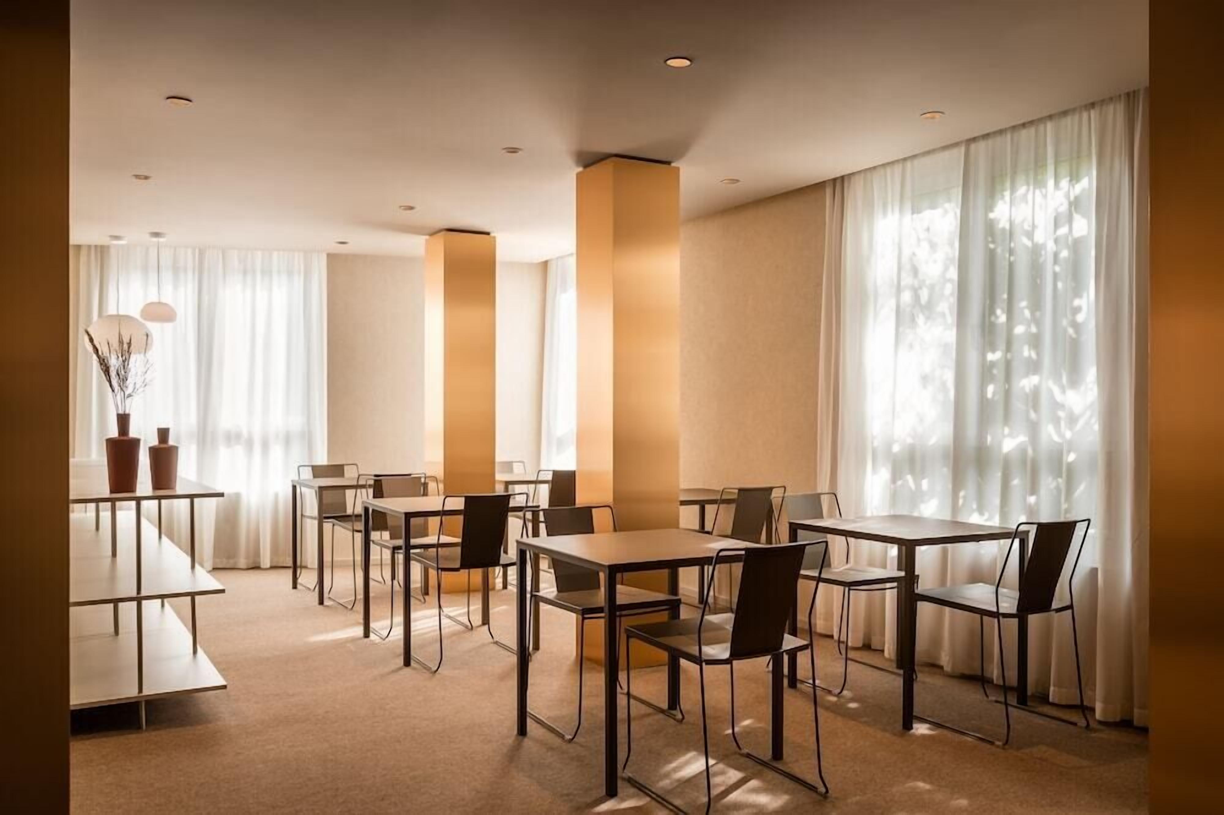 Un comedor moderno con mesas y sillas, ventanas amplias con cortinas translúcidas e iluminación empotrada en el techo.