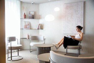 Un área de espera moderna con una mujer leyendo un libro, una mesa redonda y un cuadro abstracto grande en la pared.
