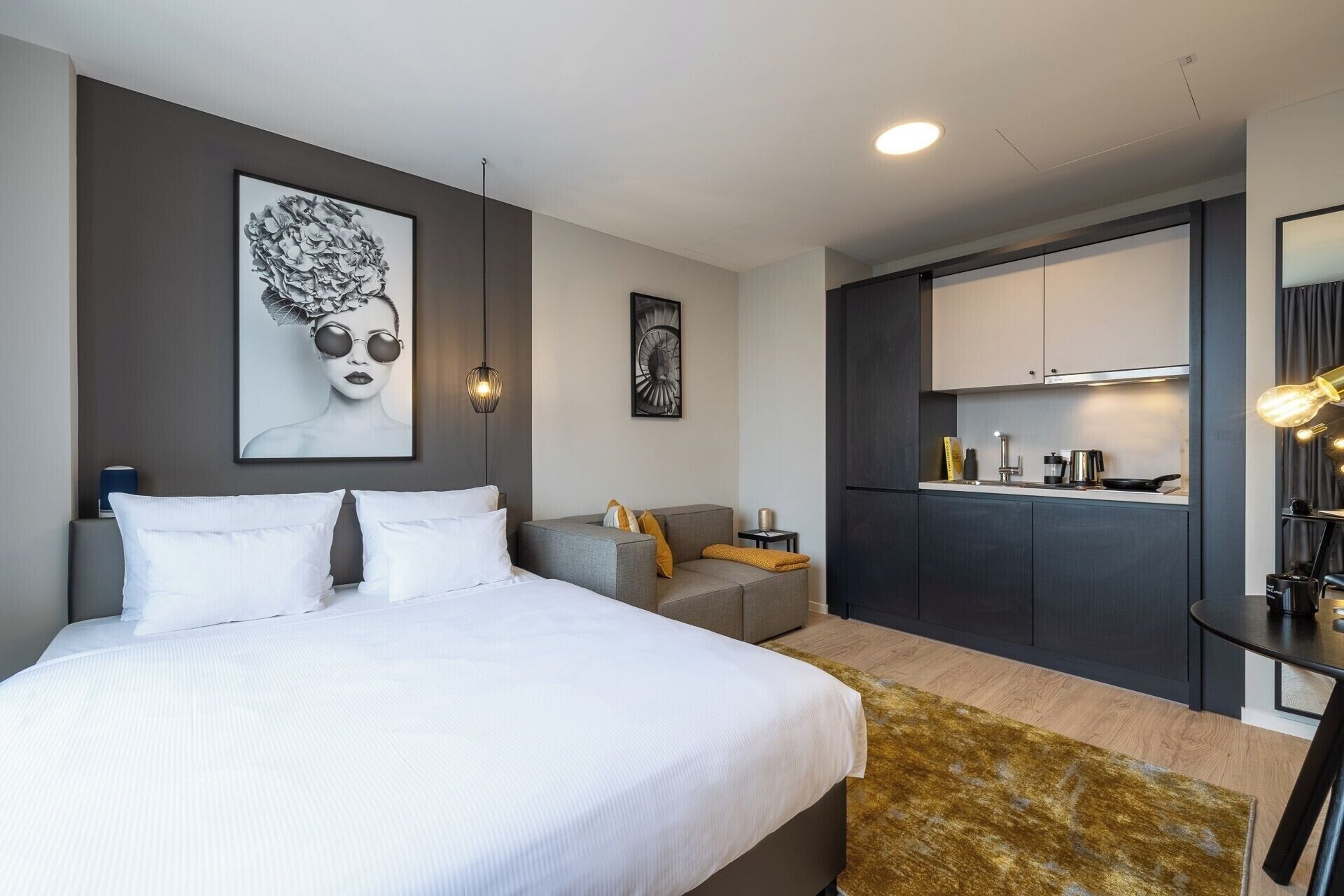 Une chambre d’hôtel moderne avec un grand lit, un canapé gris, une petite table et un coin cuisine.