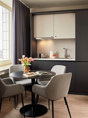 A modern kitchen with dark cabinetry, a round dining table with chairs, and a window with curtains. - Joyn Cologne (Cologne)