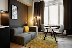 A modern living room with a grey sofa, a round table, and two chairs. There is a window with curtains, a wall-mounted light fixture, and a framed picture on the wall. - Joyn Cologne (Cologne)