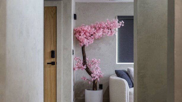 Ett rum med en dörr av trä, en vit vas med rosa blommor och en grå soffa.