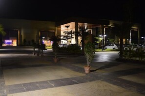 L’entrée d’un hôtel, avec un espace extérieur abrité, des plantes en pot et des voitures garées.