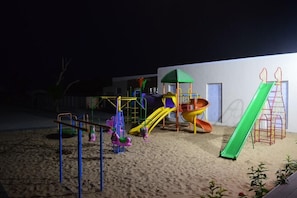 Een speeltuin met diverse speeltoestellen, zoals glijbanen en schommels, gelegen op zandbodem, met een gebouw op de achtergrond.