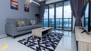 Ein modernes Wohnzimmer mit grauem Sofa, einem Holztisch und einem schwarz-weiß gemusterten Teppich. Zwei Wandklimaanlagen und ein Flachbildfernseher auf einem Holzständer sind ebenfalls vorhanden. Das Zimmer verfügt über eine große Schiebeglasdoor zum Balkon mit Blick auf die Stadt.