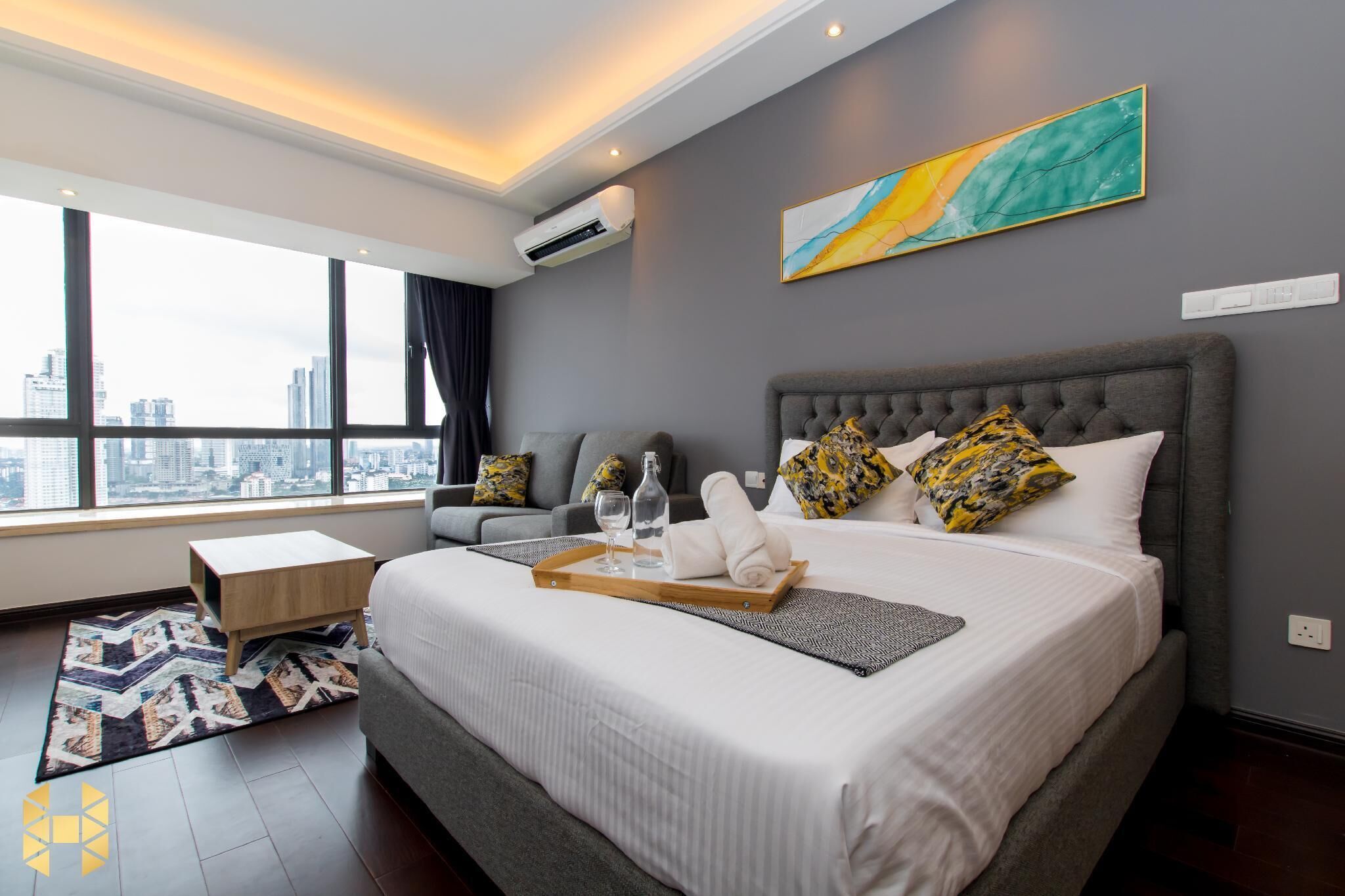 A modern hotel room with a large bed, a city view, a gray sofa, and a painting on the wall.