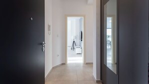 A hallway with a dark door on the left, a light-colored floor, and a mirror on the right. A door at the end of the hallway leads to a room with a tripod and a white chair. - Apartment Sang Near The Beach, Pula, Croatia (Pula)