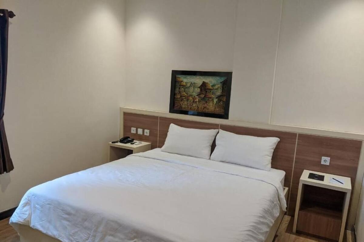 A hotel room with a large bed, a nightstand with a phone, a wall-mounted painting, and a wooden headboard.
