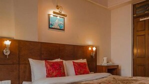 A hotel room with a wooden headboard, a bed with white and red bedding, a nightstand with a lamp, and a painting on the wall. - Hotel Shangri-la Regency (Darjeeling)