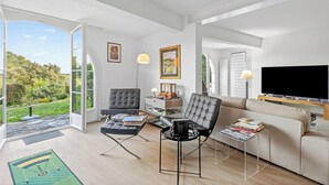 Een ruime woonkamer met een groot raam, een bank, twee fauteuils, een salontafel en een flatscreen televisie.