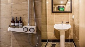Une salle de bain avec un lavabo blanc, un miroir et une douche dotée d’un panneau de commande. Trois bouteilles sont posées sur une étagère au-dessus du lavabo.