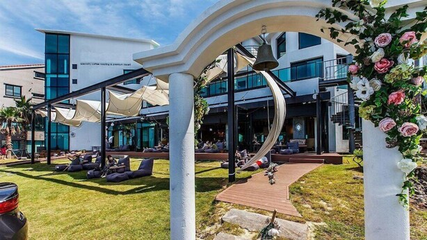 Un espace extérieur moderne, aménagé avec des fauteuils confortables et une terrasse en bois, le tout entouré d’une structure blanche agrémentée d’une cloche et de fleurs.