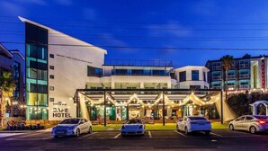 Un hôtel moderne avec un café appelé « CUTIE PIE » et une enseigne indiquant « CAFE HOTEL ». Plusieurs voitures sont garées devant.