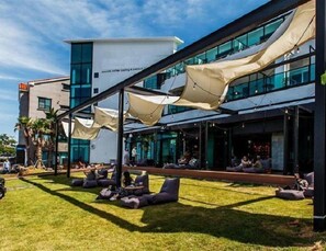 An outdoor seating area with lounge chairs and umbrellas, a modern building with large windows, and a clear sky. - Aewol Cutiepie Hotel (Jeju City)