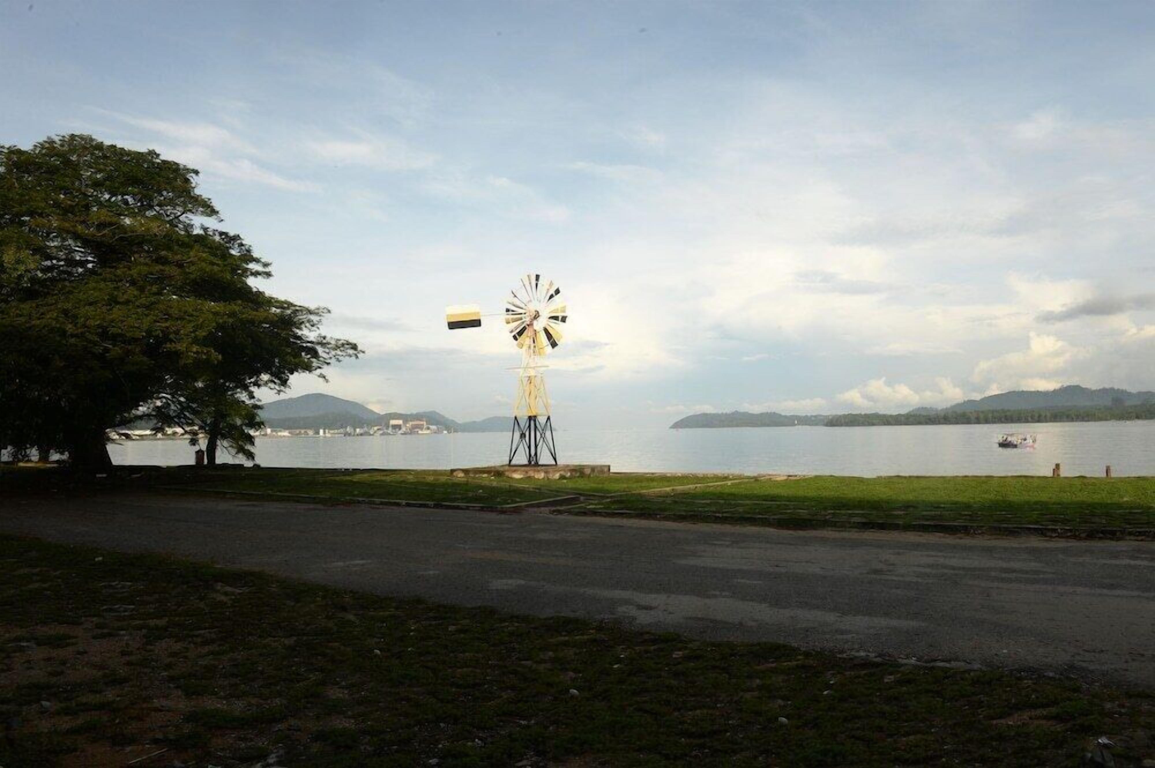 Eine Windmühle am See, im Vordergrund eine Straße und im Hintergrund Berge.