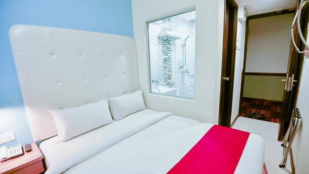 A hotel room with a large bed, a nightstand with a phone and a book, a window with a view of a building, and a bathroom with a shower and toilet. - Lumut Suites (Lumut)