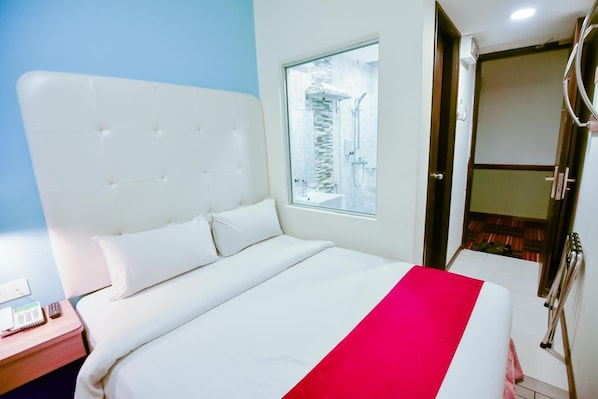 Une chambre d’hôtel avec un grand lit, une table de chevet avec un téléphone et un livre, une fenêtre donnant sur un immeuble, et une salle de bain avec une douche et des toilettes.