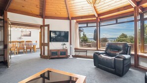 A spacious living room with a wooden ceiling, a leather sofa, a TV, and a view of the outdoors. - Aquarius home with vintage aesthetic and Lakeview (Rotorua)
