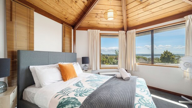 A bedroom with a bed, a bedside table, a nightstand, a window with curtains, and a view of the outdoors. - Aquarius home with vintage aesthetic and Lakeview (Rotorua)