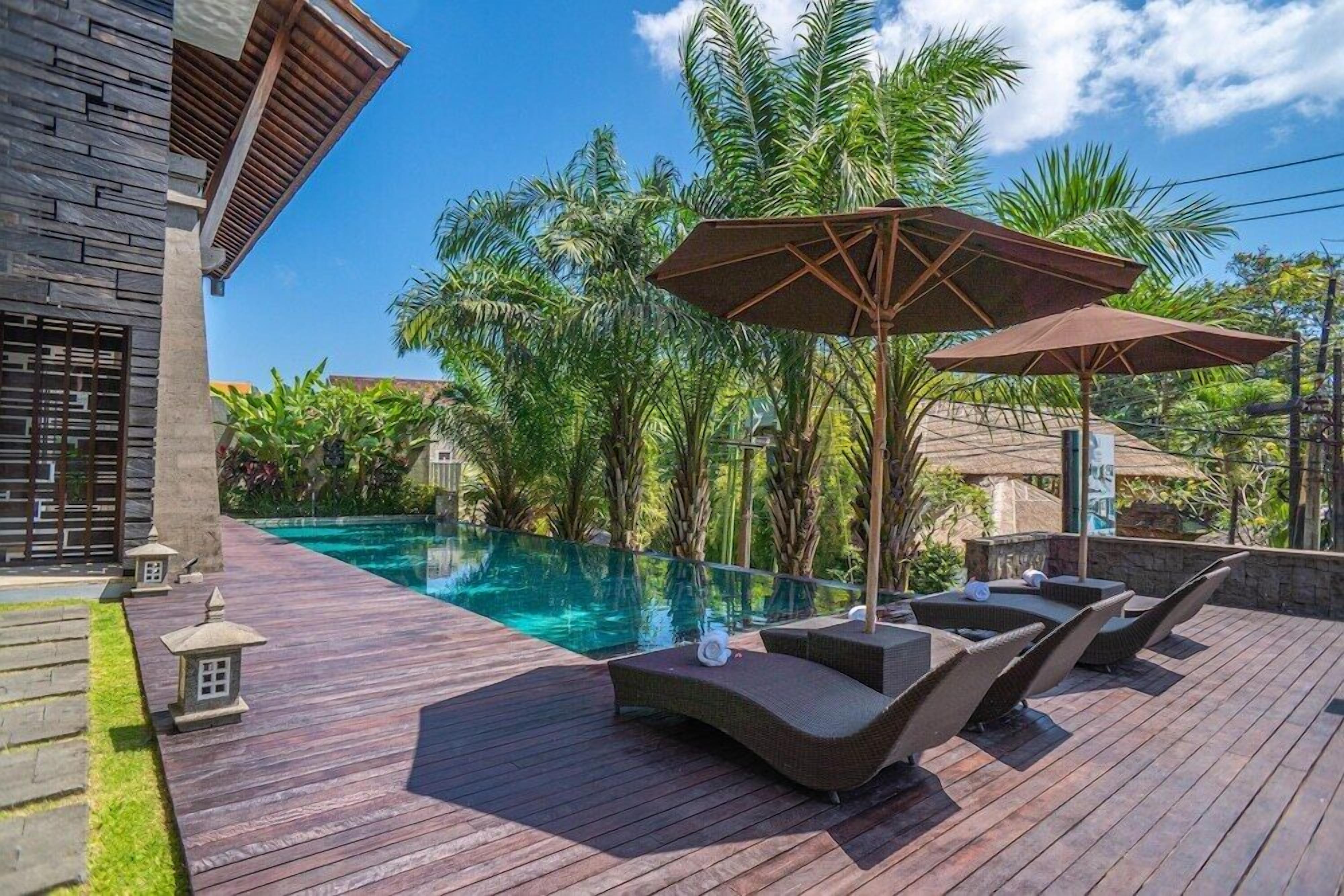 A wooden deck with lounge chairs and umbrellas, overlooking a clear pool surrounded by lush greenery.