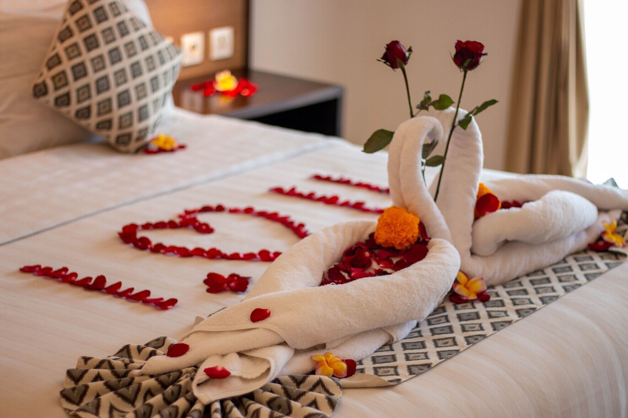 Un lit parfaitement fait, agrémenté d’une serviette pliée en forme de cygne, de pétales de rose et d’une seule rose rouge.
