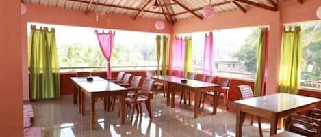 A dining area with multiple tables and chairs, large windows with curtains, and a high ceiling with wooden beams.