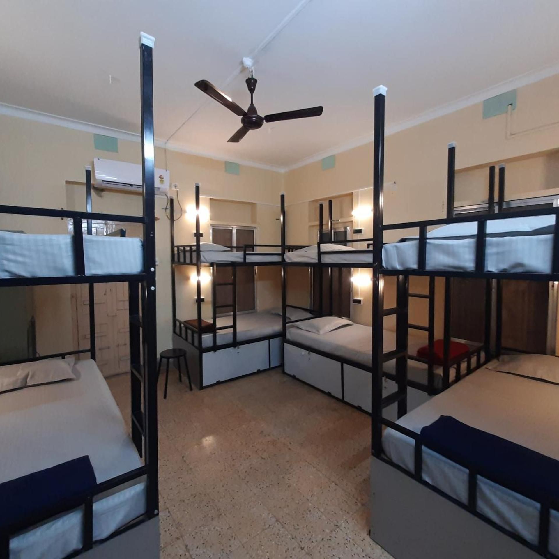 A dormitory room with bunk beds, a ceiling fan, and a kitchenette in the background.