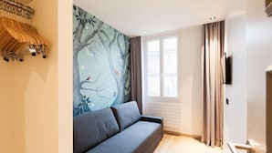 Une pièce avec un canapé gris, une fenêtre avec des rideaux, un mur orné d’une fresque représentant un arbre et un porte-manteau.