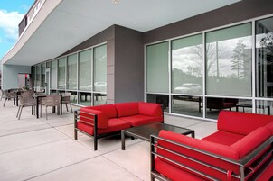 Un área de estar al aire libre con sofás rojos y mesas, ventanas amplias y una fachada de edificio moderna.