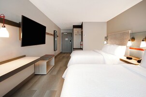 Habitación de hotel con dos camas, un televisor de pantalla plana montado en la pared, un escritorio con una silla y una mesita de noche con lámpara.