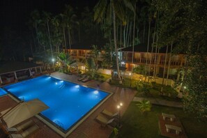 Vista noturna de um hotel com piscina, espreguiçadeiras e palmeiras.