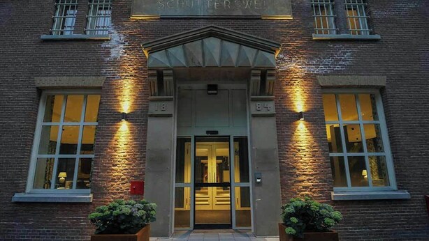 A brick building with a sign reading 'SCHUTTERSWEI' at the entrance, large windows, and two planters with greenery. - The Fallon Hotel Alkmaar (Alkmaar)
