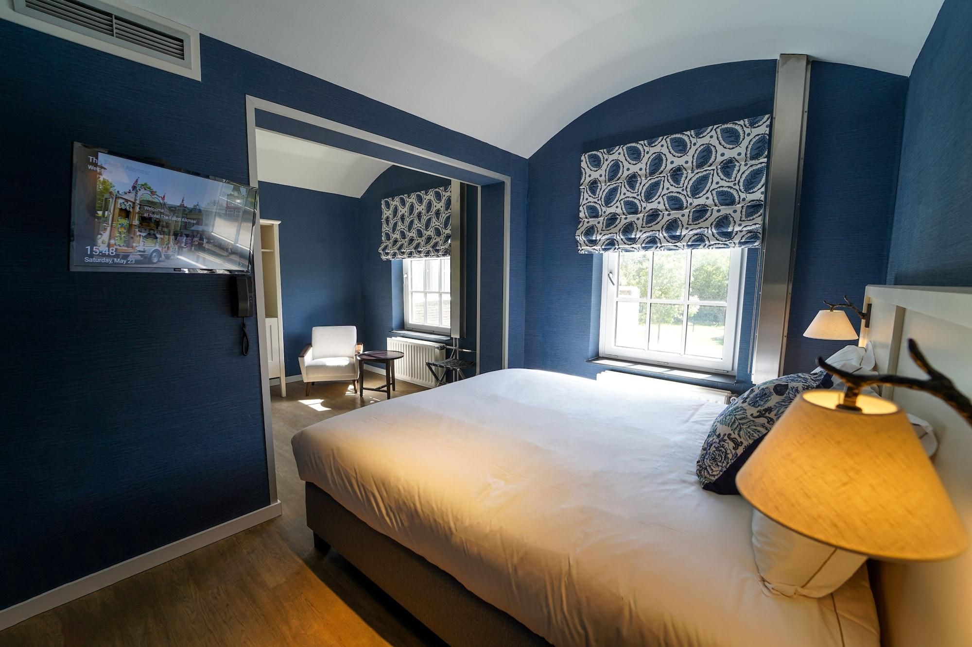 A bedroom with a bed, a nightstand, a lamp, a window with patterned curtains, a mirror, and a view of the outside.