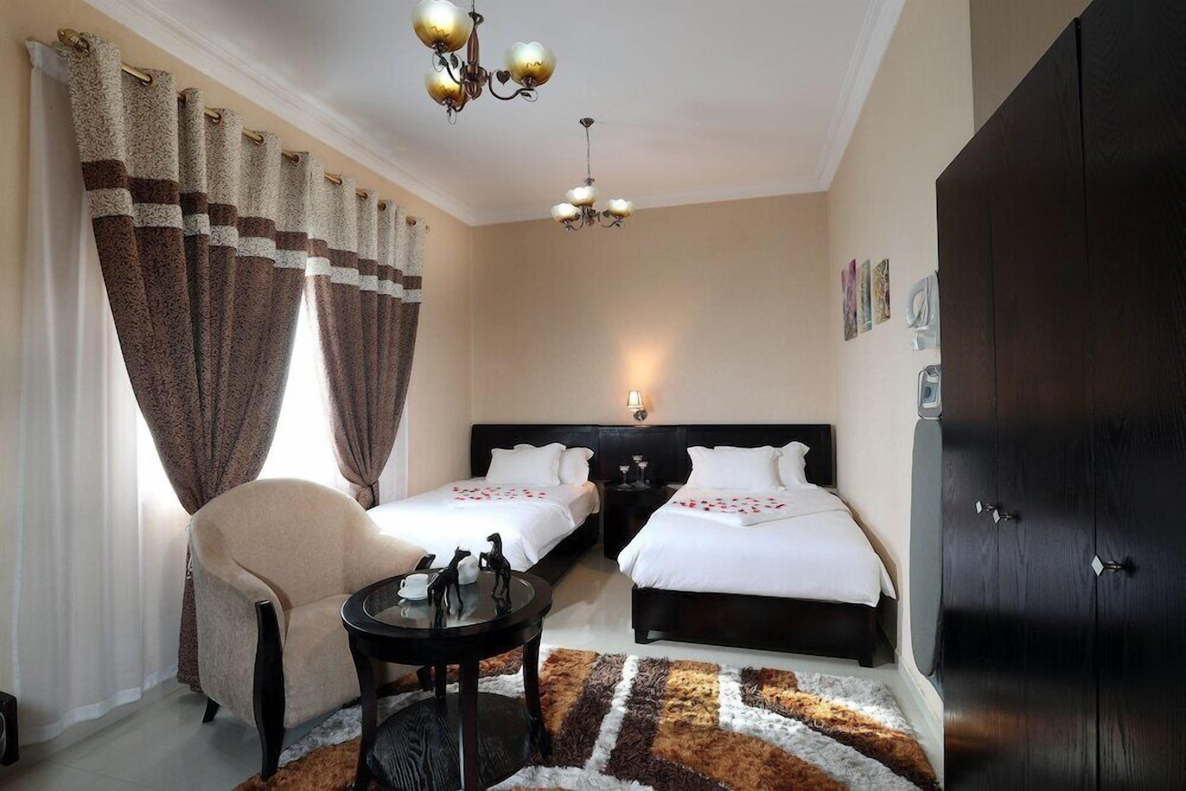 A hotel room with two beds, a sitting area with a chair and a small table, a chandelier, and a wardrobe.