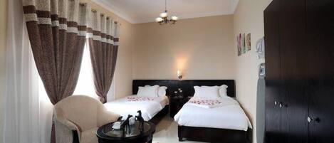 A hotel room with two beds, a sitting area with a chair and a small table, a chandelier, and a wardrobe.