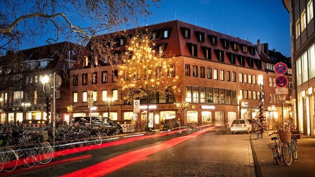 A multi-story building with a brick facade, multiple windows, and a tree with lights in front. - H.ostel Munster (Münster)
