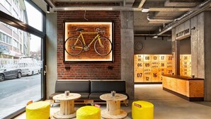 A modern interior with a large window, a bicycle artwork, a reception desk, and a seating area with round tables and yellow ottomans. - H.ostel Munster (Münster)