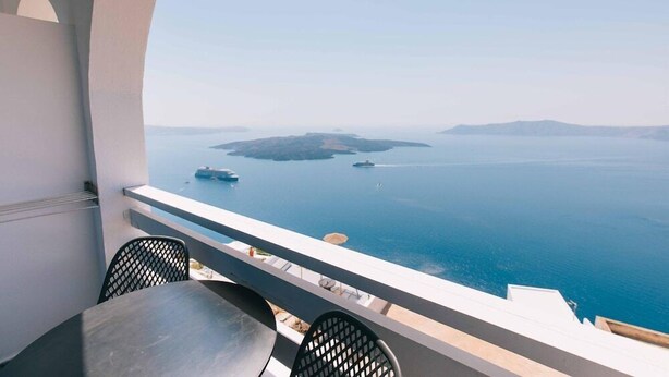 A balcony with a table and chairs overlooking a calm sea with boats and islands. - Kafieris View (Santorini)