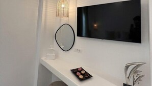 A modern living room with a flat-screen TV mounted on a white wall, a pendant light, a round mirror, a small white table with a plate of cookies, and a decorative metal sculpture. - Kafieris View (Santorini)