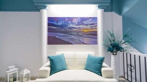 A room with a white sofa, teal cushions, a painting of a sunset over the ocean, and a white vase with blue foliage. - Kafieris View (Santorini)