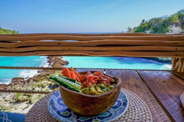 Ein Holztisch mit einer gewebten Tischmatte und einer Salat-Schüssel mit Blick auf die Küste.