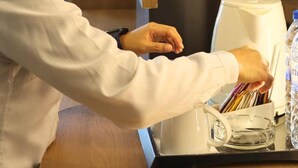 A person seated at a table with a coffee machine, a glass of water, and a bottle of water.