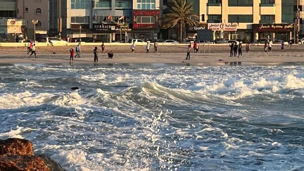 A beachfront hotel with multiple floors, a sandy beach, and waves crashing onto the shore.