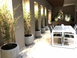 A modern outdoor dining area with a white table and chairs, surrounded by large potted plants and a stone pathway.