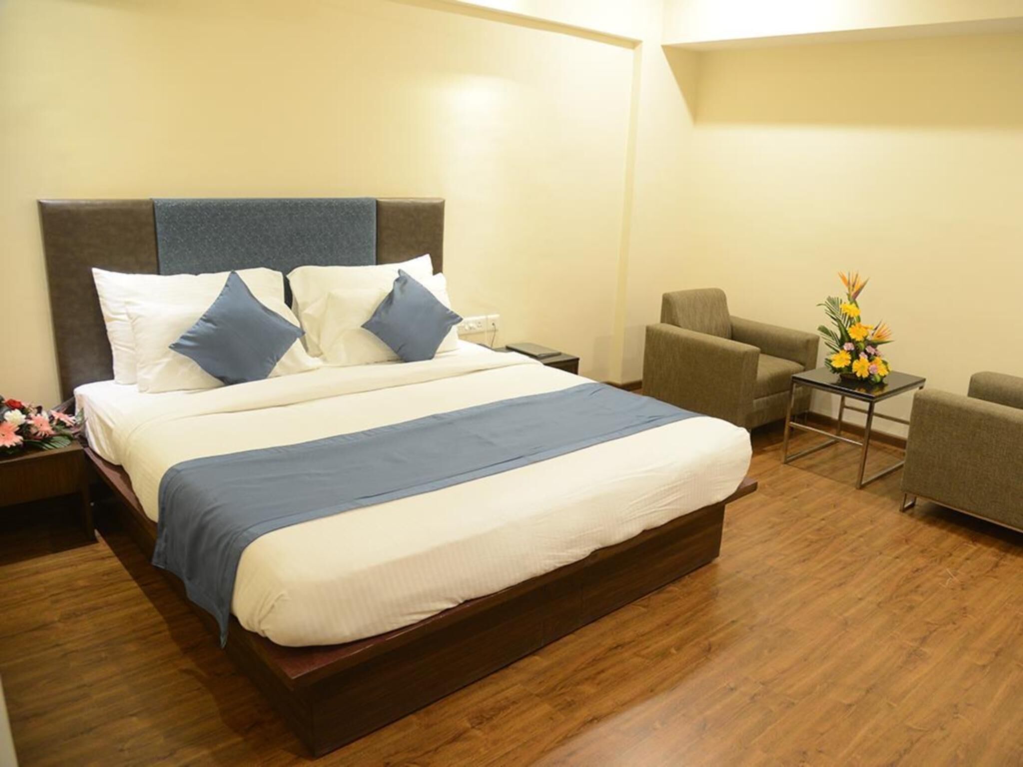 Habitación de hotel con una cama grande, una mesita de noche, una silla, una pequeña mesa con un jarrón de flores y piso de madera.