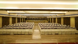 A conference room with rows of white chairs and tables, a stage at the front, and a high ceiling with lighting fixtures.