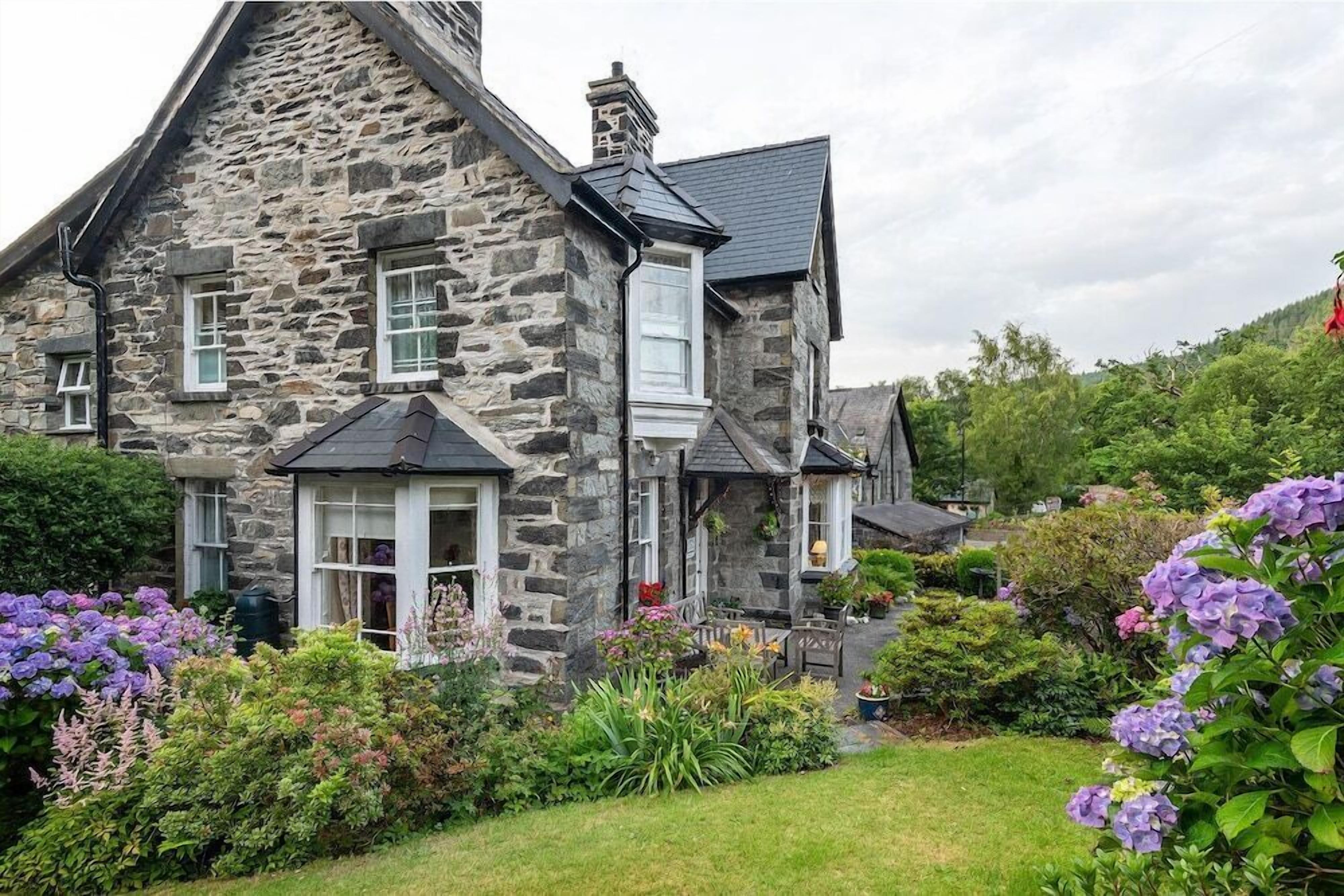A stone house with a steeply pitched roof, multiple windows, and a well-maintained garden with various plants and flowers.