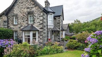 A stone house with a steeply pitched roof, multiple windows, and a well-maintained garden with various plants and flowers.