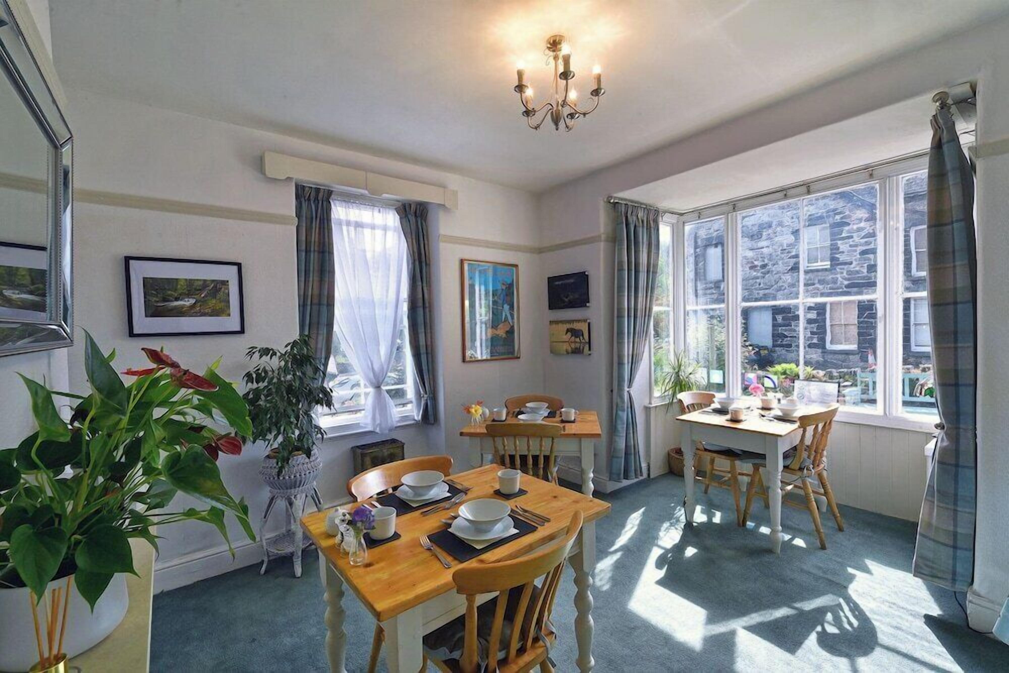 A dining area with wooden tables and chairs, a chandelier, and large windows with curtains.
